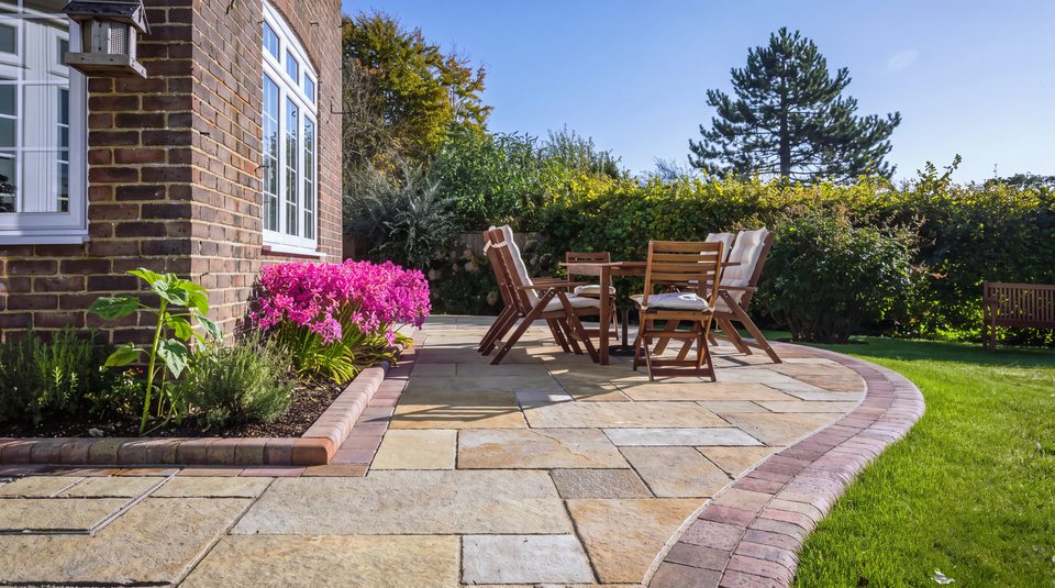 A natural stone patio in full sun beside a brick house, with a striped lawn beyond